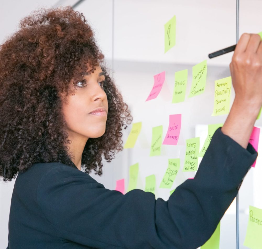 beautiful-young-businesswoman-writing-on-sticker-with-marker-concentrated-professional-curly-female-manager-sharing-idea-for-project-and-making-note-min